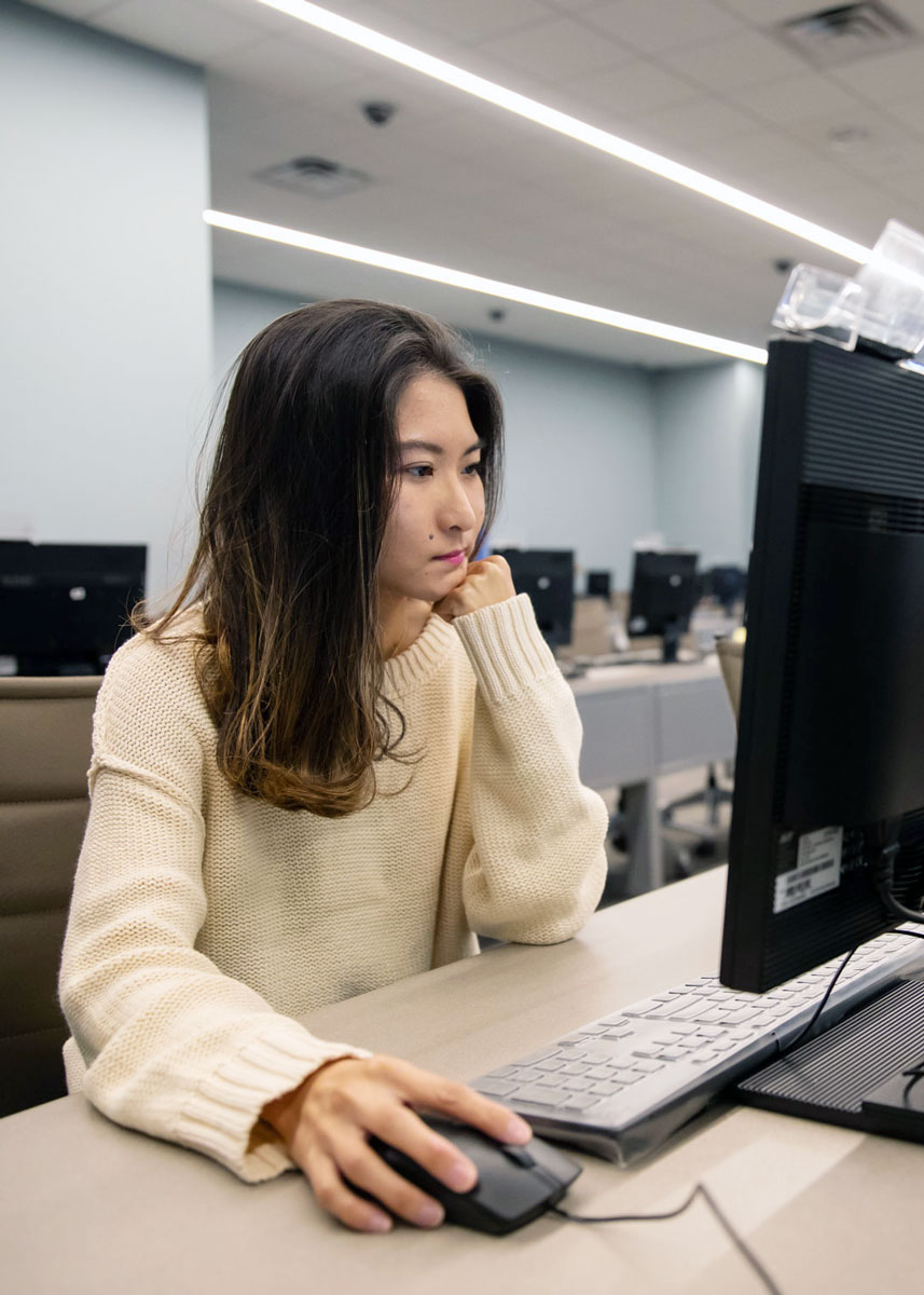 A woman sits at a computer, looking at the screen and using a mouse.