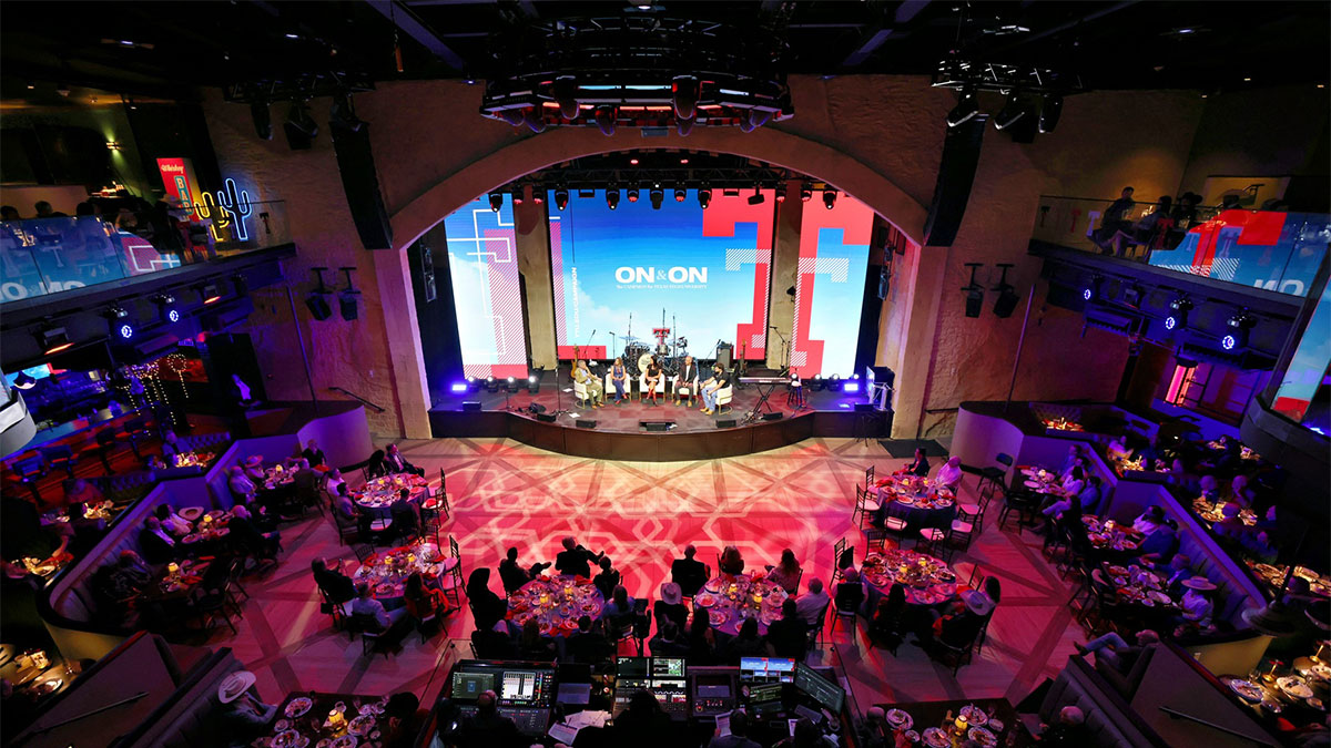Aerial view of a formal event venue with round dinner tables filled with guests. A stage at the front of the room features a panel of five speakers seated in chairs beneath large LED screens displaying the ON&ON: The Campaign for Texas Tech University logo. The room is lit in red and purple with the Texas Tech double T logo projected on the floor.
