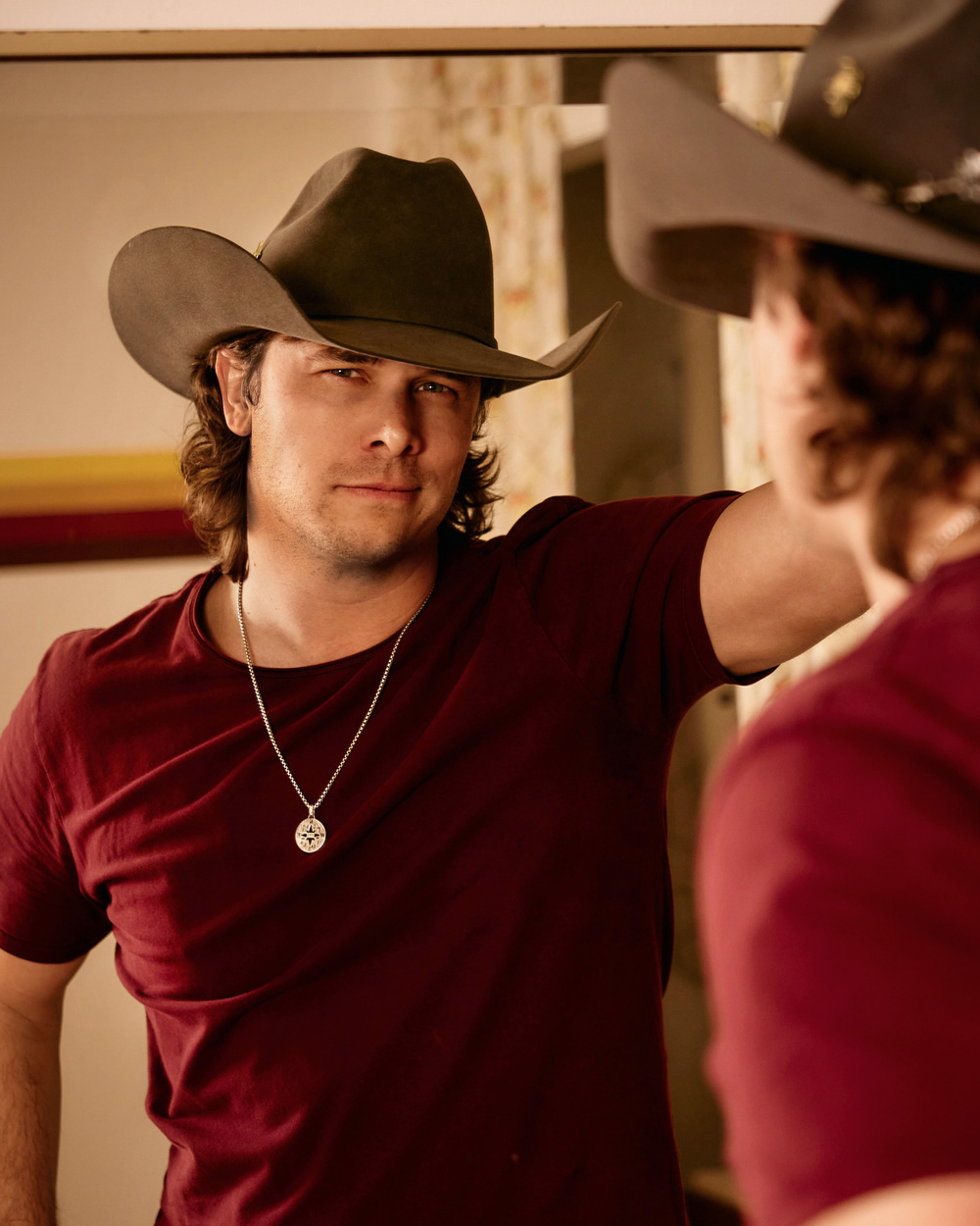 Randall King in a cowboy hat and maroon shirt looks at his reflection in a mirror.