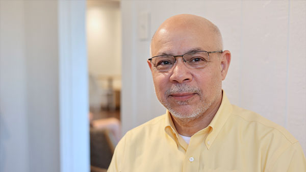 Dr. Abidi wears glasses and a light yellow button-down shirt while posing for a portrait indoors. A softly blurred doorway and room are visible behind him.