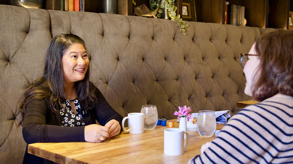 Narissra Punyanunt-Carter and Lucy Greenberg share a conversation during a meal at a restaurant, with bowls of food, mugs and condiment bottles on the table