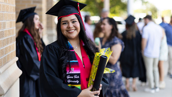Jennifer Mendoza Trejo smiles in her Texas Tech graduation regalia while holding her diploma and flowers