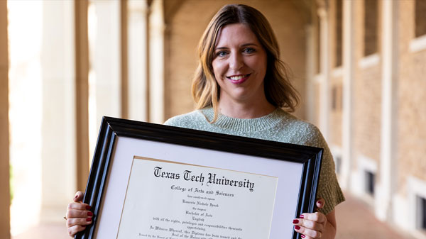 A woman stands in a campus archway holding a framed Texas Tech University diploma and smiling toward the camera.