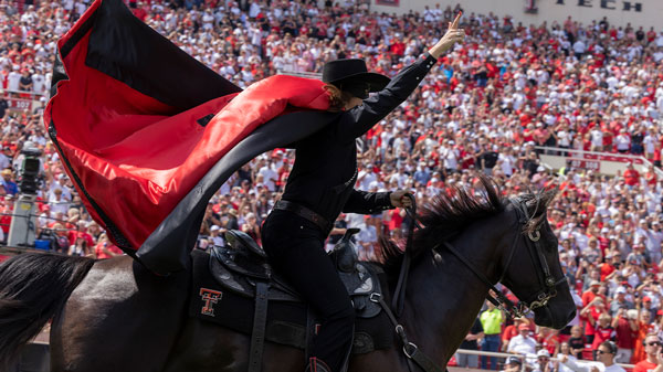 The Masked Rider races across the field before a Texas Tech football game, waving a large red-and-black cape as a packed stadium crowd cheers