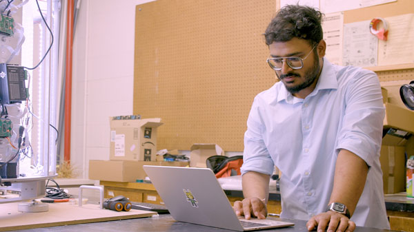 Mayank Bangari works on a laptop beside a prototype scanning device in an engineering laboratory filled with electronics and testing equipment