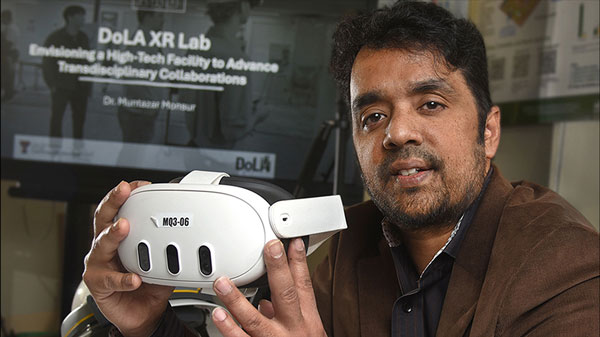 Man holds a virtual reality headset in front of a screen displaying information about an XR lab and interdisciplinary collaboration