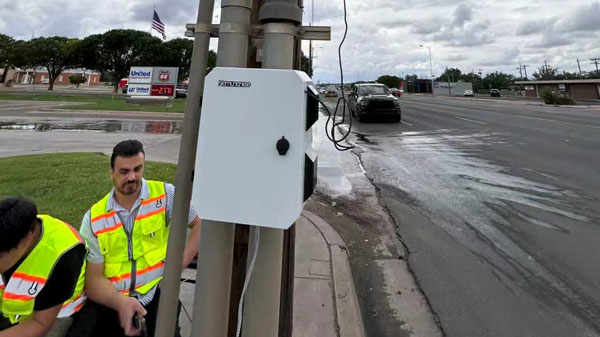 Workers in safety vests install equipment on a utility pole beside a roadway