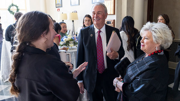 A man in a suit and red tie, Ben Strickling, speaks with his wife Roxane and another woman during a crowded indoor reception, with guests, tables and floral arrangements in the background.