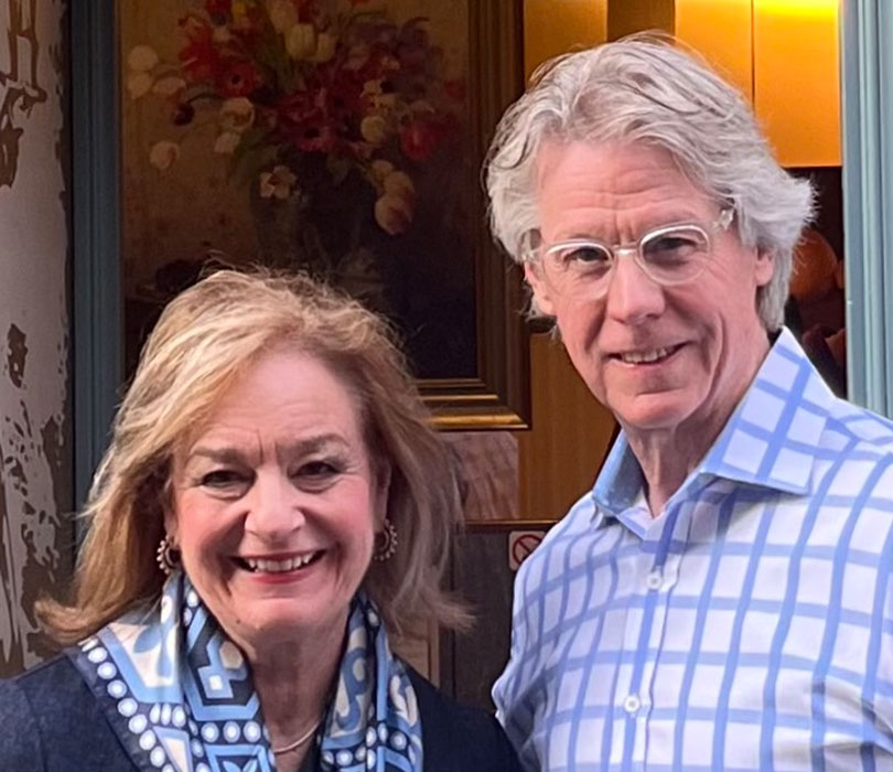 A smiling David and Lorna Griffin stand side by side outside a building entrance, Lorna is wearing a patterned blue scarf and David is wearing glasses and a blue checkered shirt.