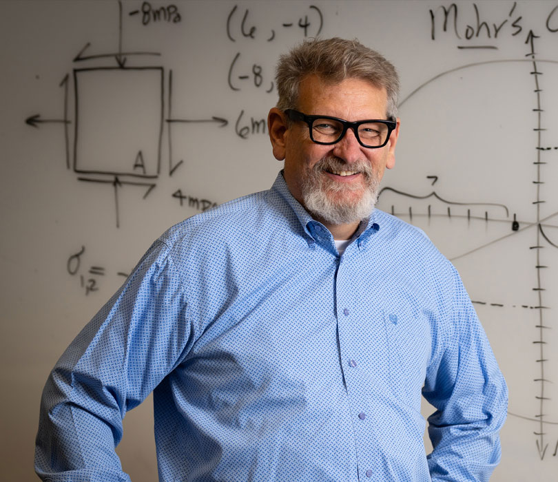 A smiling man with glasses and a gray beard stands with his hands on his hips in front of a whiteboard covered with engineering equations and diagrams, including Mohr's circle.