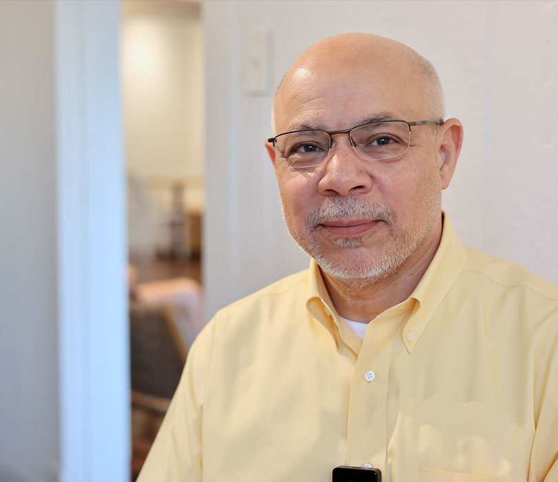 Dr. Abidi wears glasses and a light yellow button-down shirt while posing for a portrait indoors. A softly blurred doorway and room are visible behind him.