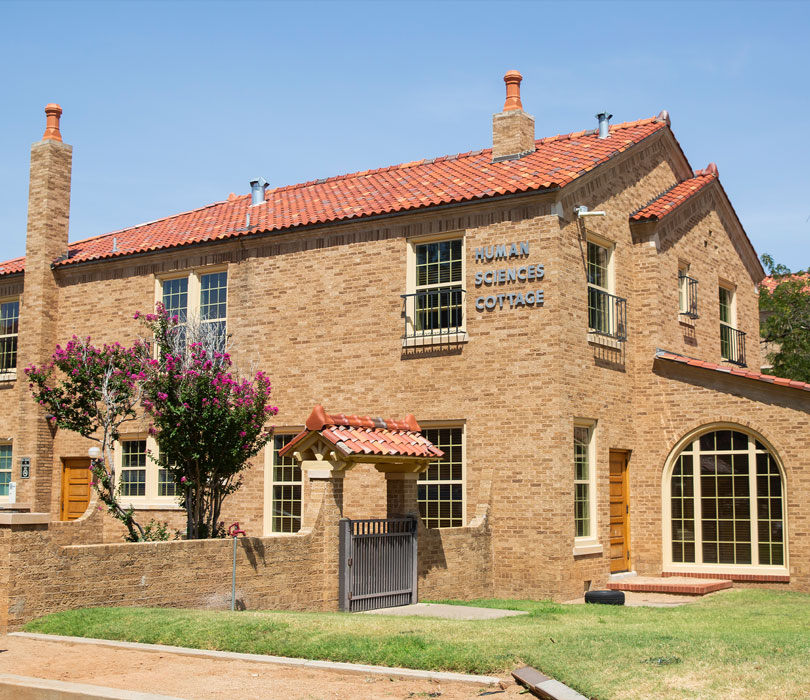 The Human Sciences Cottage, a two-story brick building with a red tile roof, arched windows and exterior signage, stands on the Texas Tech campus with a small courtyard and flowering tree in front.