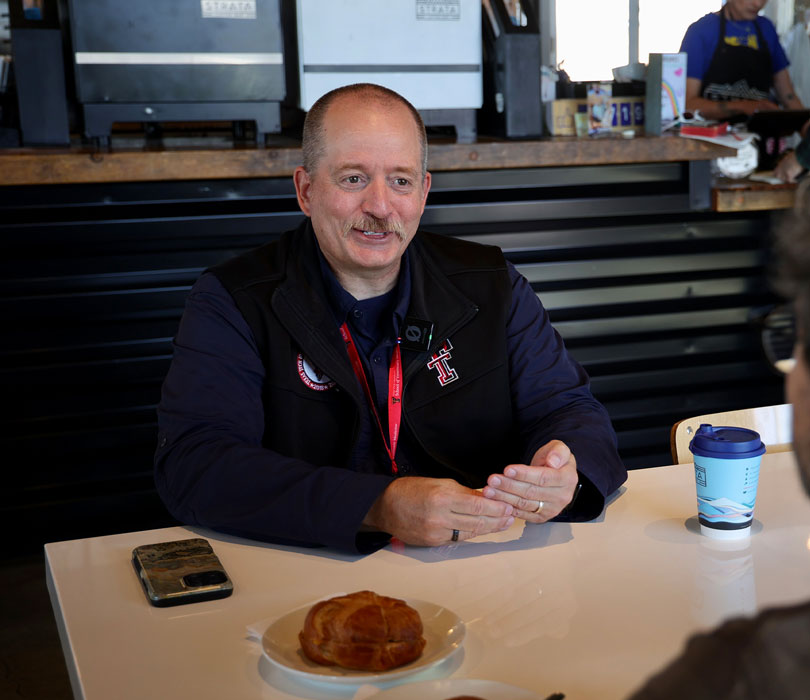 Man sits at a café table with coffee and a pastry, speaking to someone off-camera.