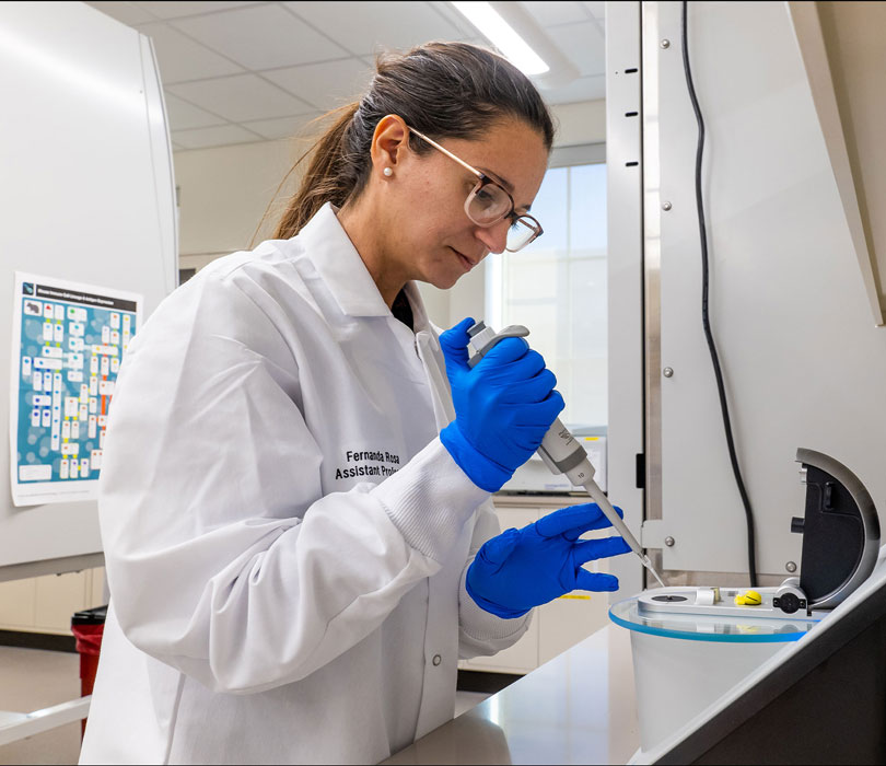 Researcher in a white lab coat and blue gloves uses a pipette while working at a lab station.