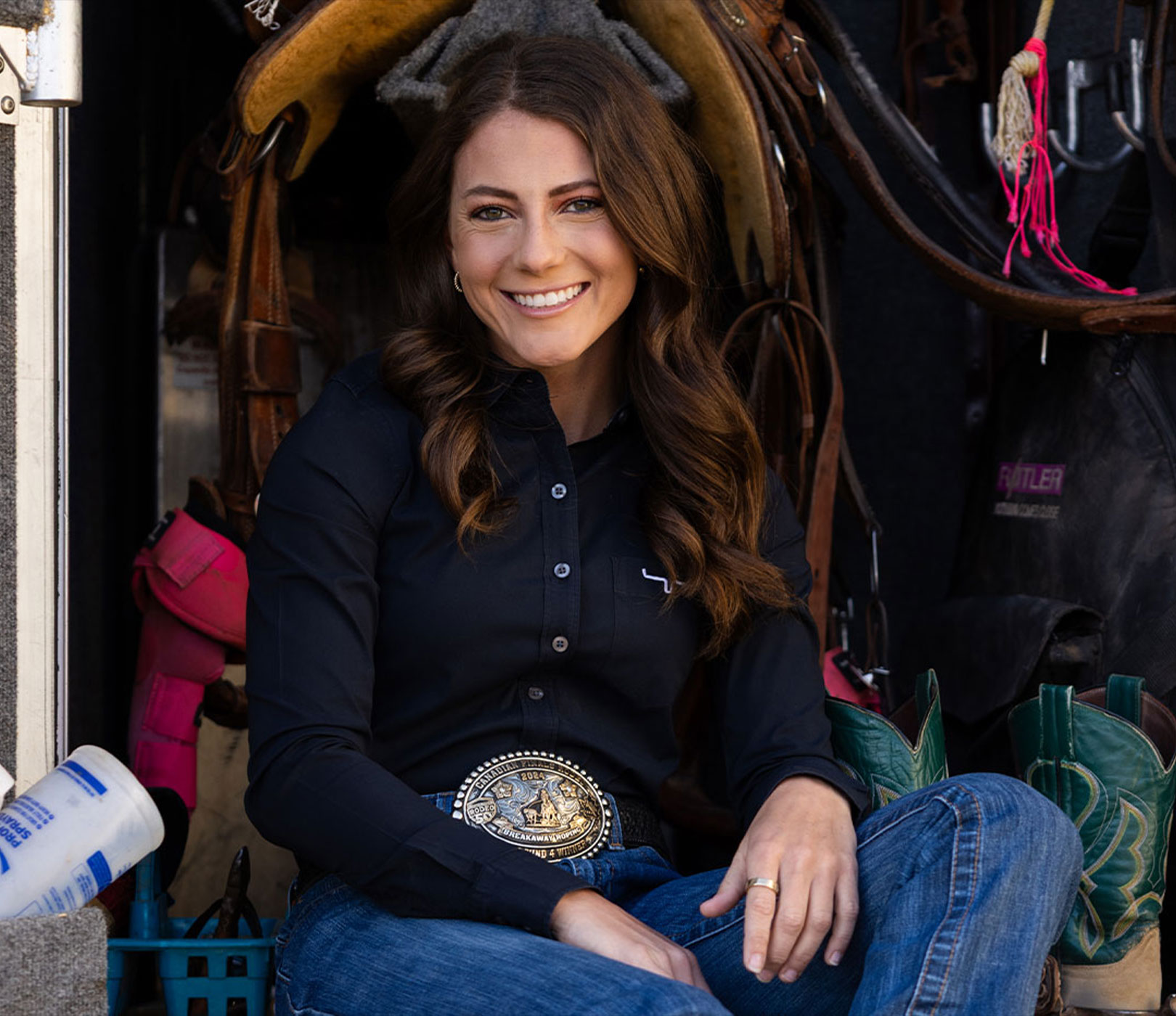 Jessi in western attire sits smiling among saddles and riding gear.