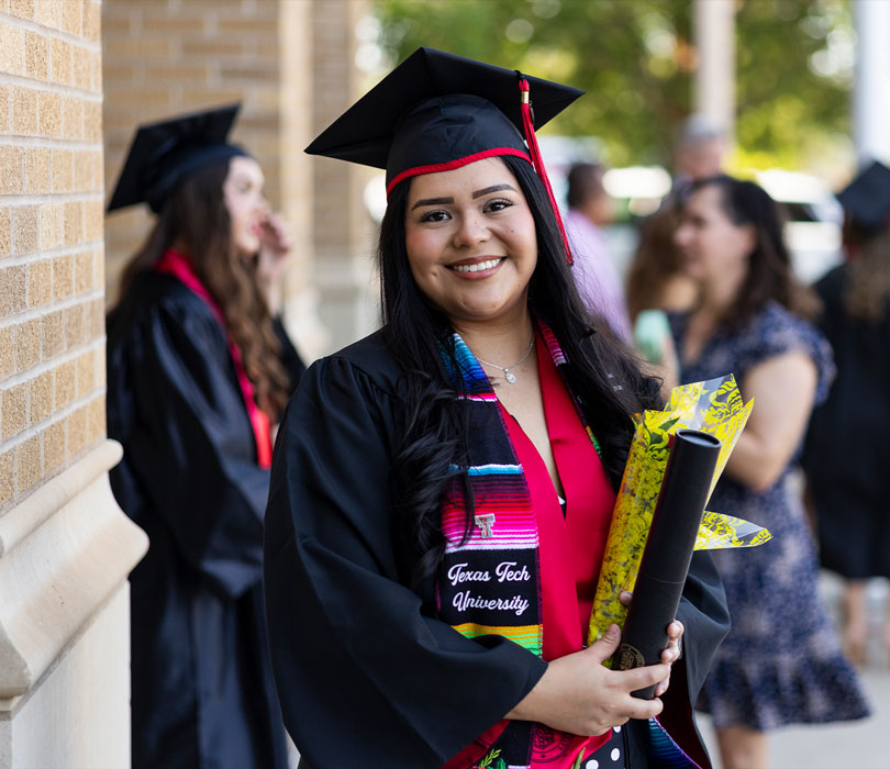 Jennifer Mendoza Trejo smiles in her Texas Tech graduation regalia while holding her diploma and flowers.