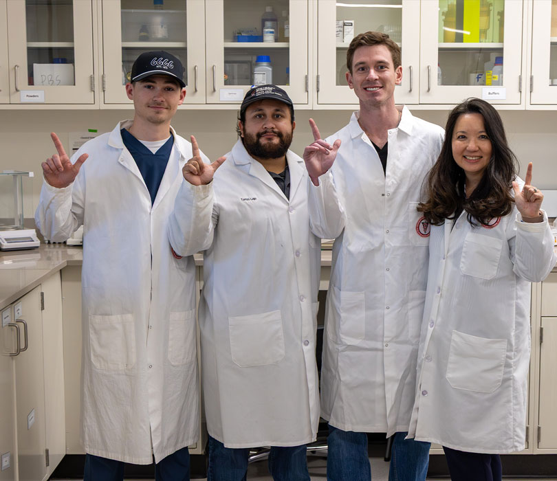Four members of a Texas Tech research team pose in a laboratory wearing white lab coats and making “Guns Up” hand signs.