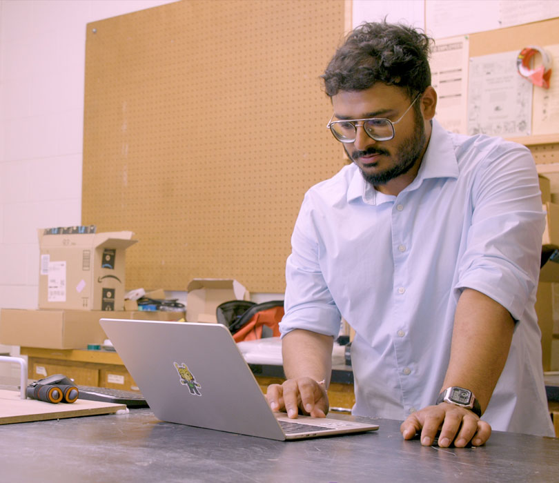 Mayank Bangari works on a laptop beside a prototype scanning device in an engineering laboratory filled with electronics and testing equipment.