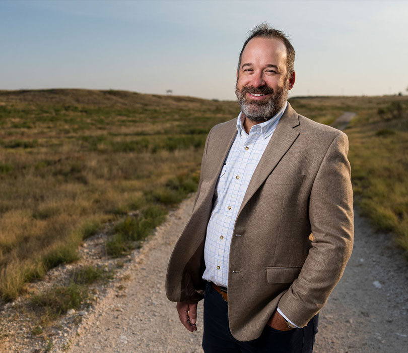 A smiling man in a blazer stands in a sunlit West Texas grassland, surrounded by rolling prairie beneath a clear blue sky.
