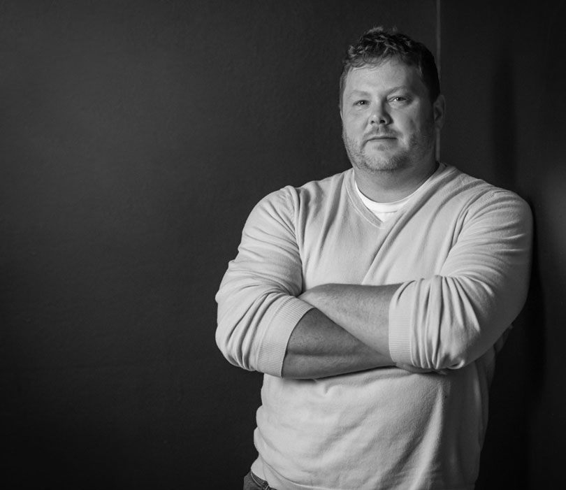 A black-and-white portrait of Daniel Pruske with arms crossed against a dark background.