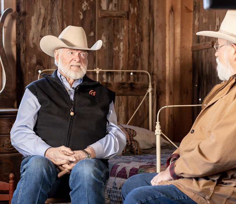 Two white-bearded men wearing cowboy hats sit facing each other in conversation inside a rustic wooden room at the National Ranching Heritage Center.