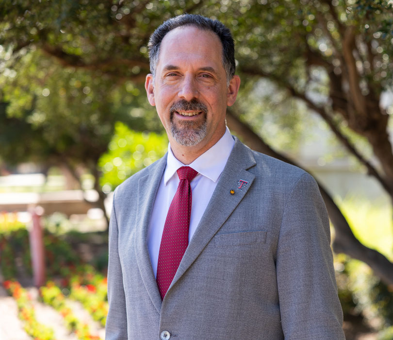 James Reecy smiles while standing outdoors beneath mature trees on campus. He wears a gray suit jacket, white dress shirt and red tie, with Texas Tech lapel pins on his jacket. The softly blurred background features landscaped flower beds, walkways and campus greenery in bright daylight.