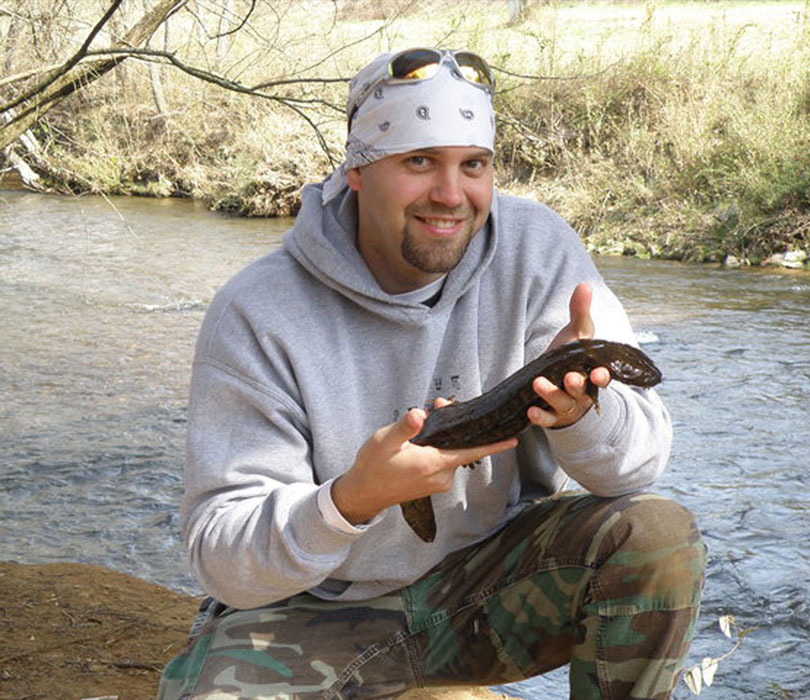 Rod Williams kneels beside a stream while holding a hellbender salamander during fieldwork.