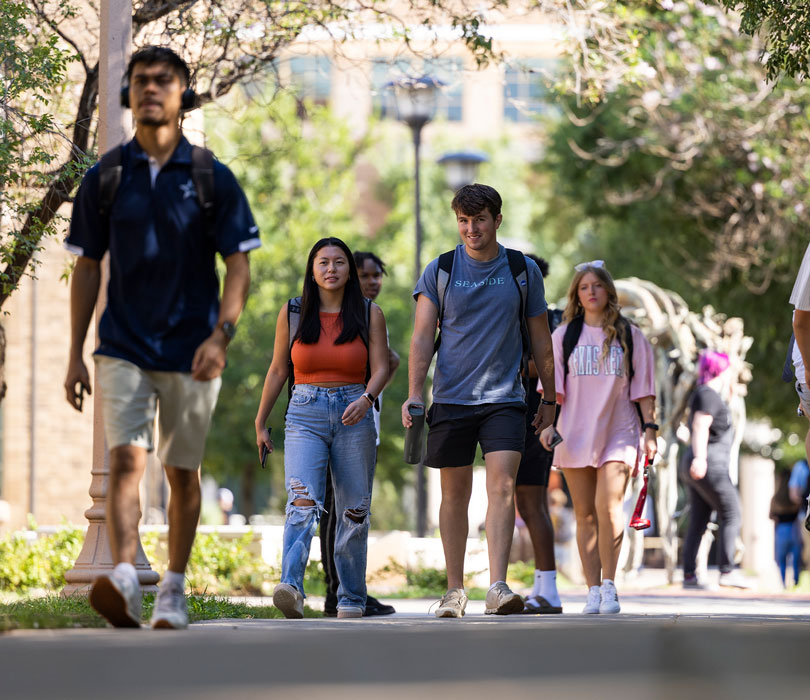 Texas Tech students wearing backpacks walk along a tree-lined campus sidewalk between classes.