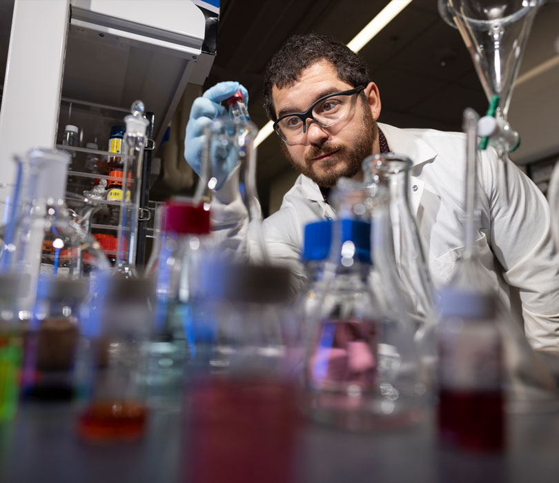 A researcher wearing a lab coat, safety glasses and gloves handles a small vial while surrounded by beakers, flasks and other glassware filled with colorful liquids.