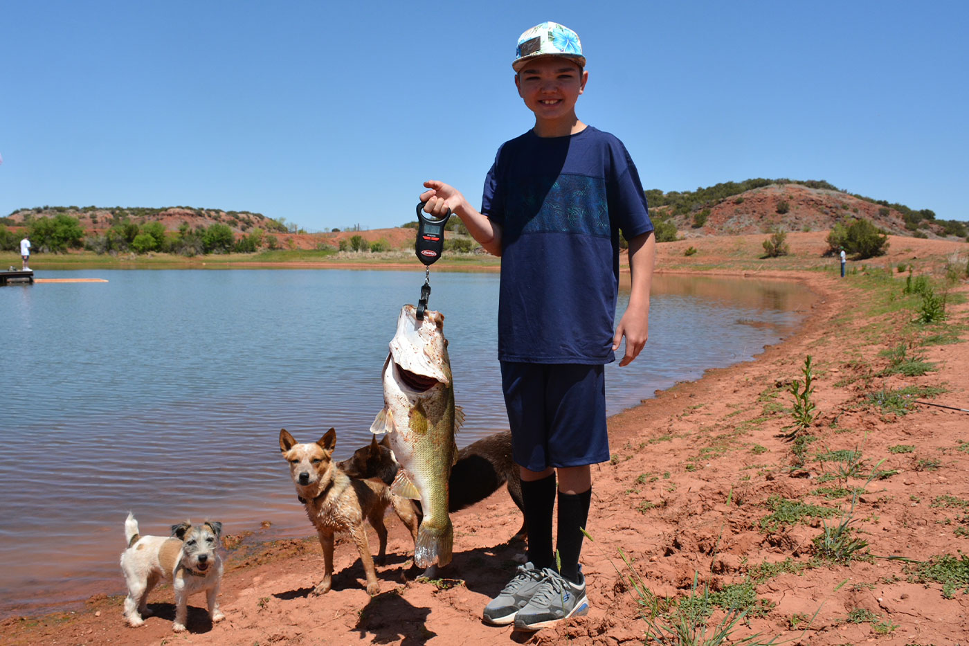 Jett just caught a fish. He is holding it while standing next to a pond alongside his dogs. 