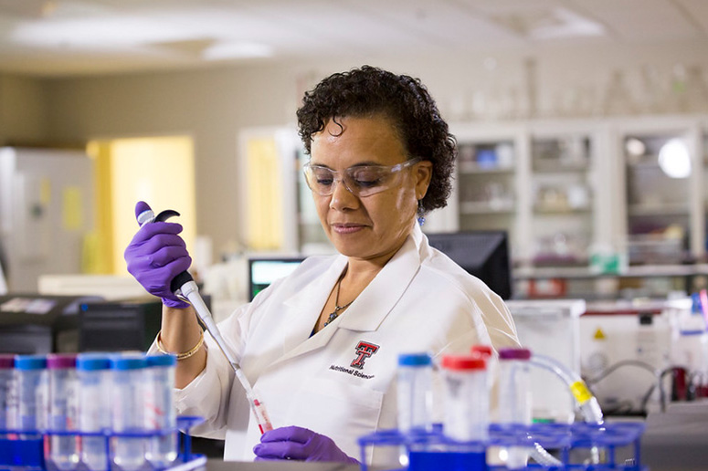 A woman in a lab coat holding a pipette and test tube, with a tray of test tubes in front of her and lab equipment in the background.
