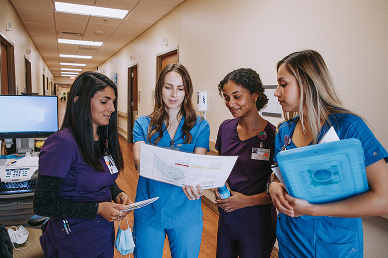 Four interns wearing scrubs looking at a printed chart while standing in a hospital hallway.